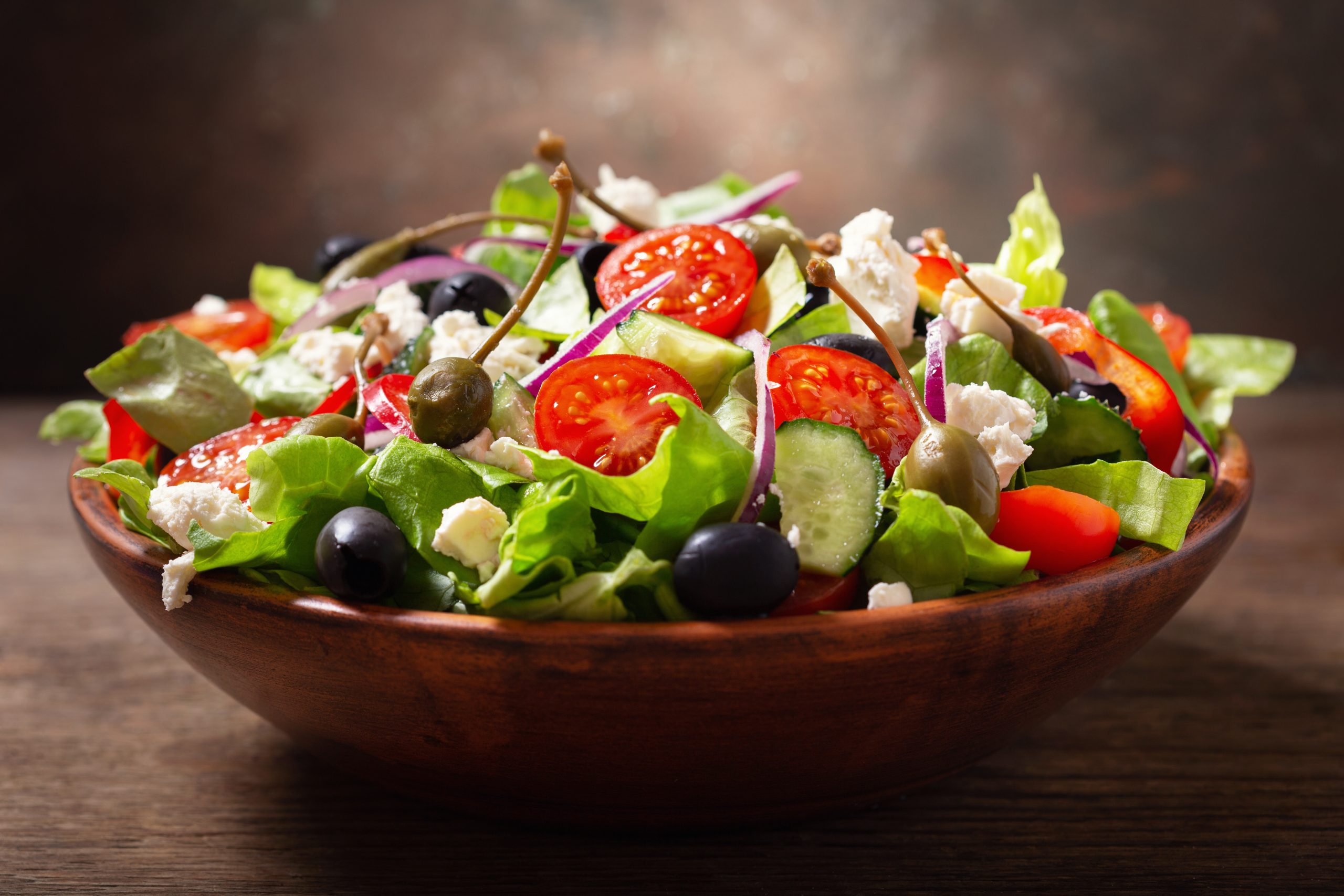 bowl of fresh salad with vegetables, feta cheese and capers on a wooden table